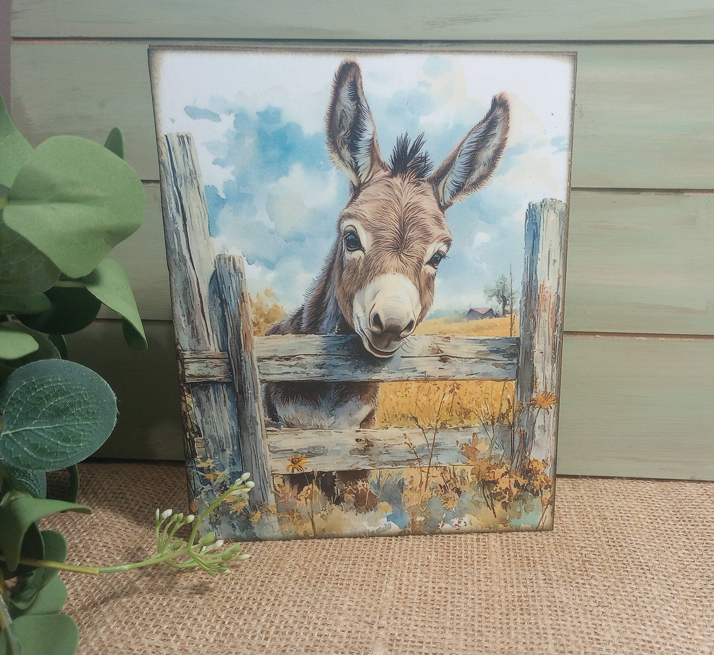 Painting of a donkey peering over a wooden fence with a natural landscape background.