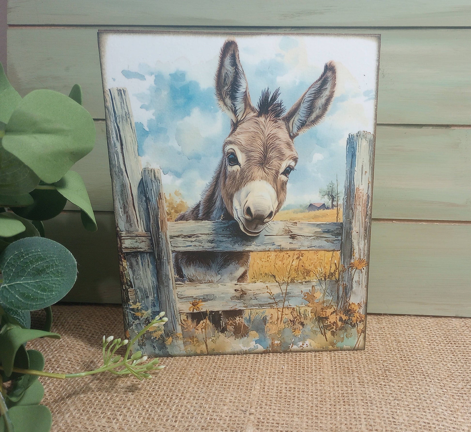 Painting of a donkey peering over a wooden fence with a natural landscape background.